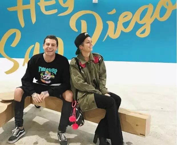 A smiling couple poses for a photo in front of a vibrant blue wall with yellow lettering that says "Hey San Diego". The man wears a black sweatshirt and ripped jeans, while the woman is in a green jacket and black leggings.