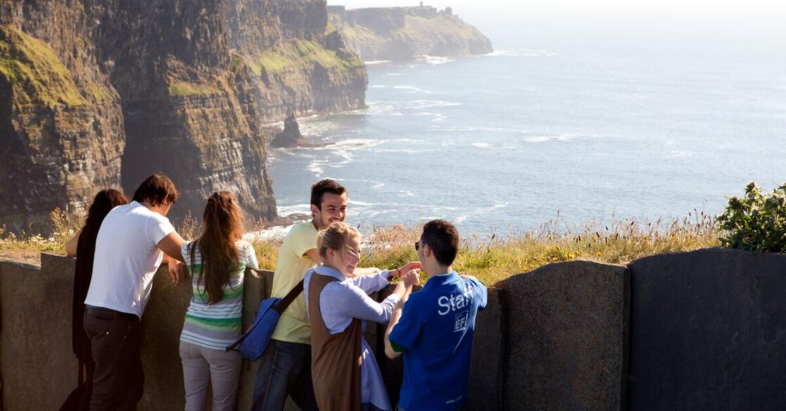 A group of tourists stands on a cliff overlooking the sea and the rocks. They appear to be enjoying the view.
