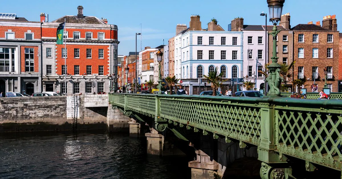 A green bridge spans a river, with buildings in the background. The architecture suggests a European city, likely Dublin, Ireland, featuring its characteristic red brick buildings and the River Liffey.