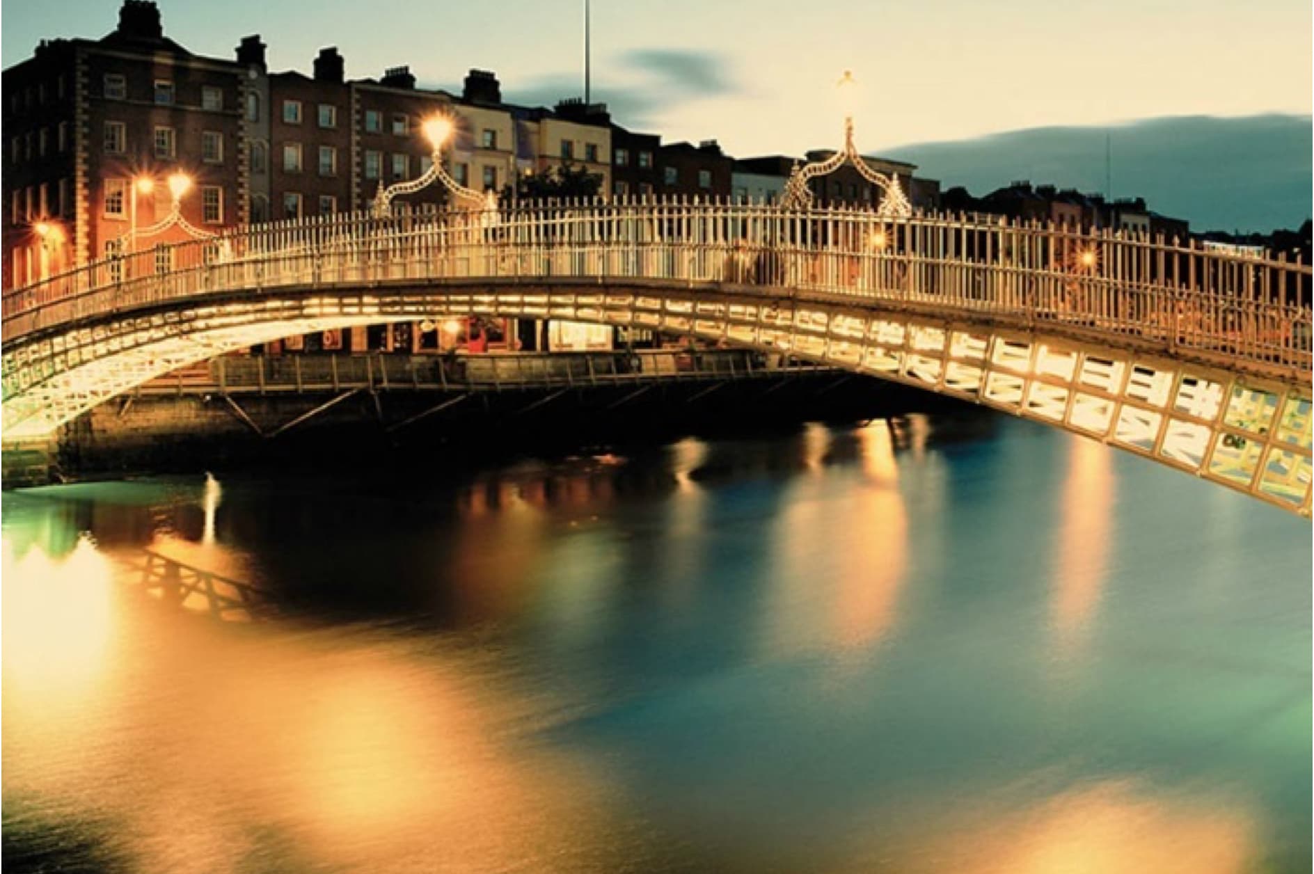 The image shows the Ha'penny Bridge in Dublin, Ireland, illuminated at dusk. The iconic pedestrian bridge spans the River Liffey, with historic buildings lining the background and their reflections shimmering on the water.