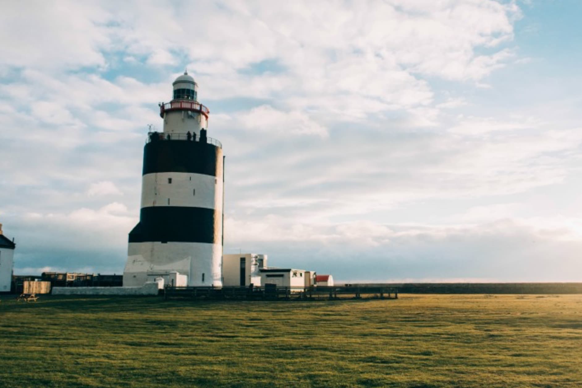 A striking black and white striped lighthouse stands on a grassy field under a dramatic sky with clouds and sunshine.