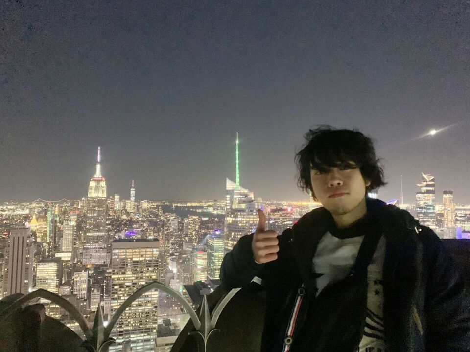 A young person gives a thumbs up while posing for a photo at night with a cityscape in the background. The city appears to be New York City, with the Empire State Building visible.