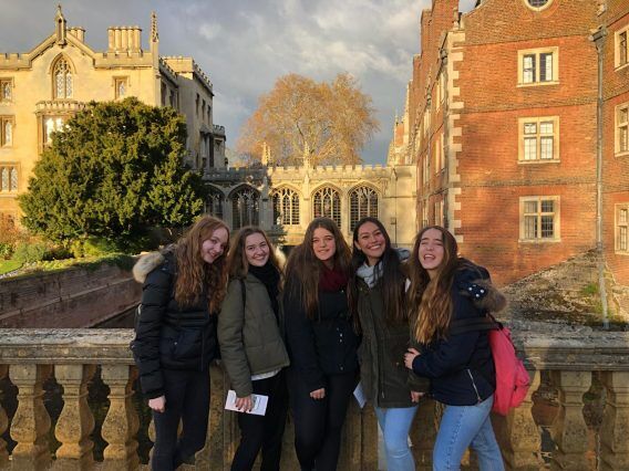 Five young women smile on a bridge in front of historic brick and stone buildings, illuminated by trees and sunshine.