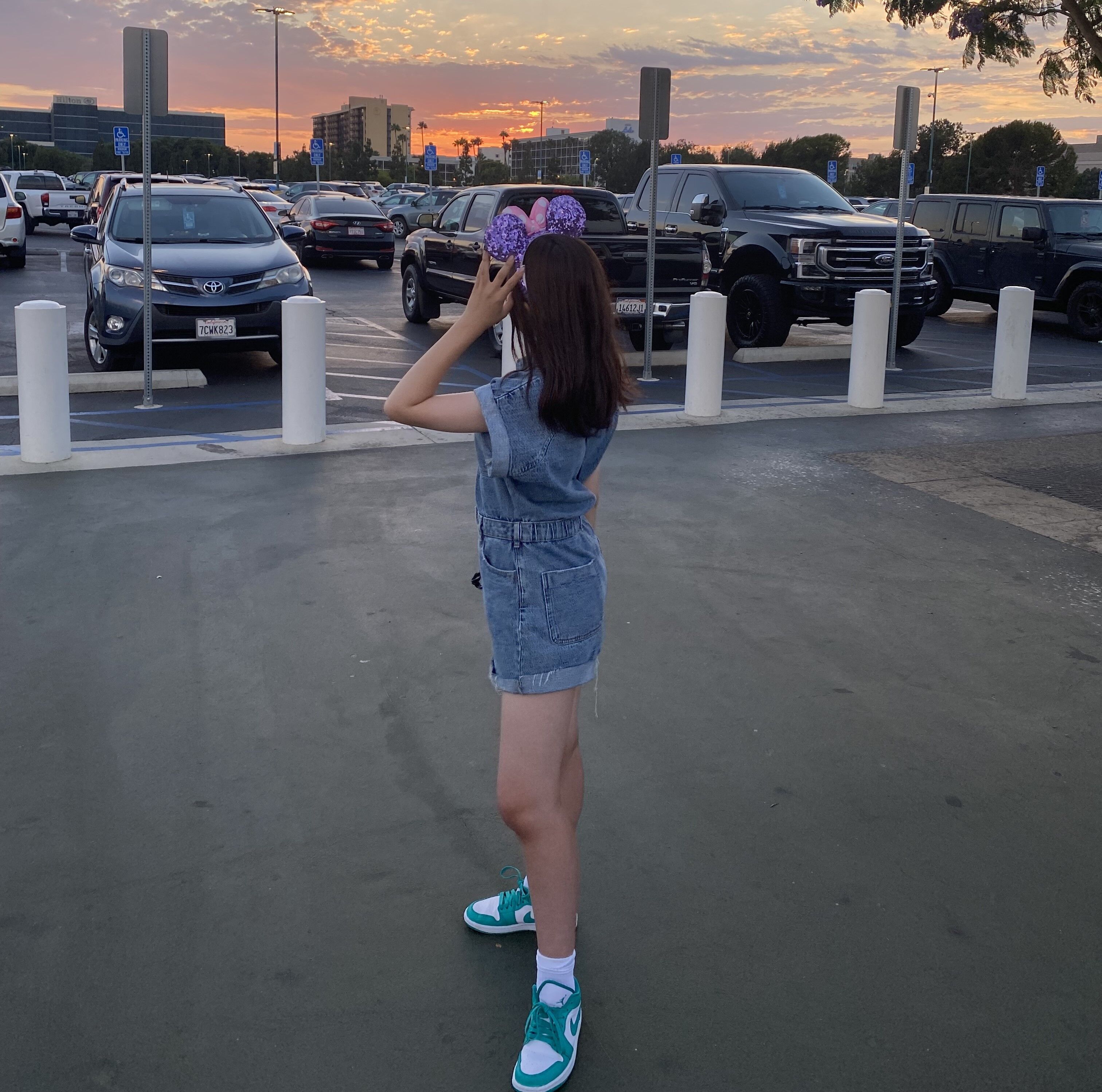 A young girl wearing Minnie Mouse ears stands in a parking lot, holding her hand up to her ears as she looks at the camera. She is wearing a denim overall and sneakers, with a sunset in the background.