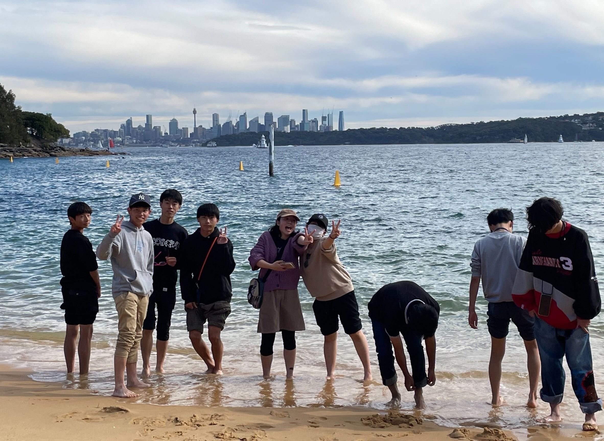 A group of young people stand on a beach, looking out at the water with a city skyline in the distance.