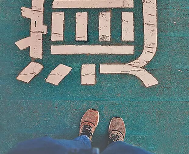 The image shows a close-up view of a person's feet standing on a teal-colored surface with a large, white, weathered Japanese kanji character painted on it. The character appears to be "道" (michi), meaning "road" or "way".
