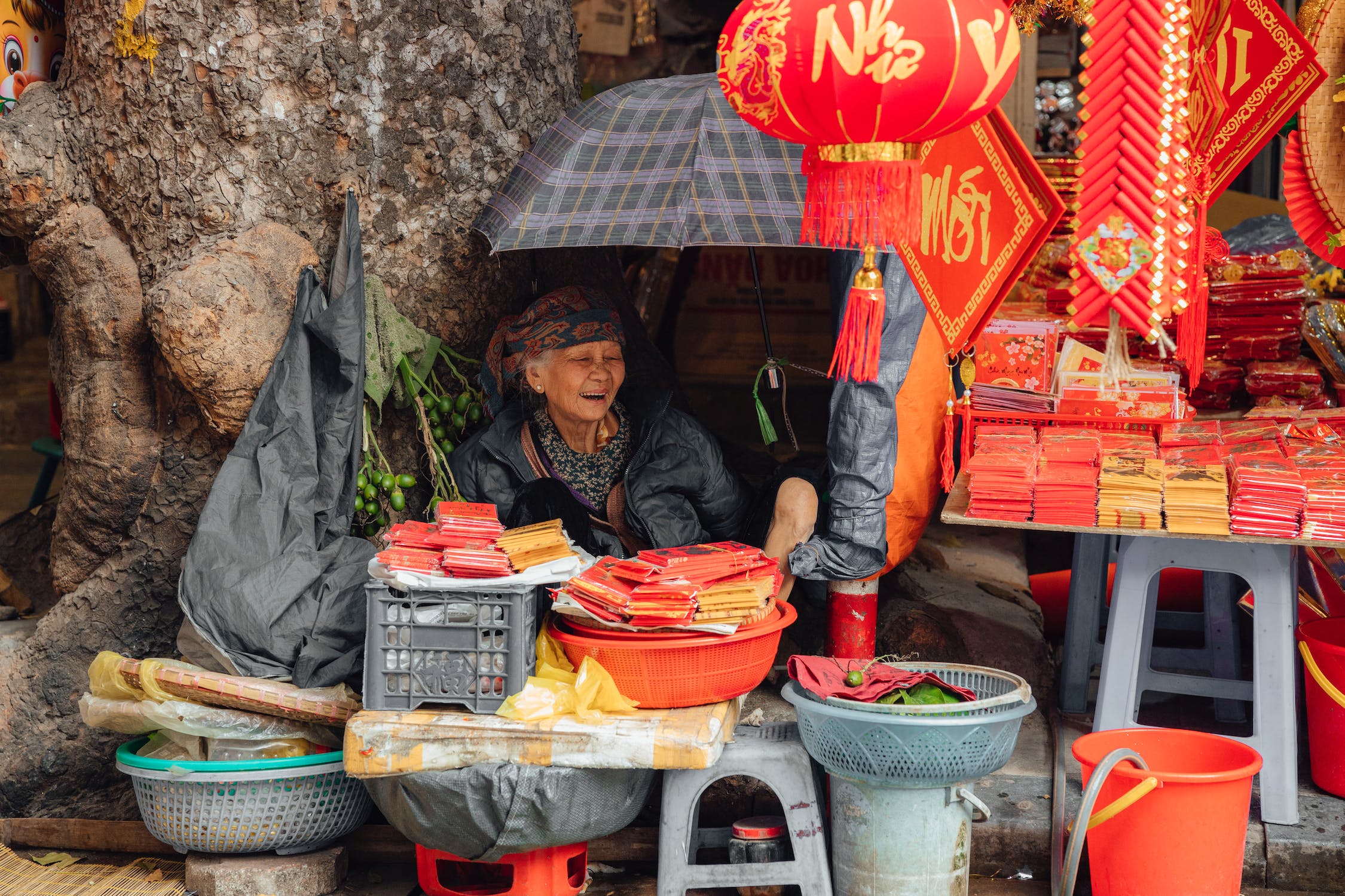 An elderly woman sits under an umbrella, selling red papers and other items at a bustling market stall.