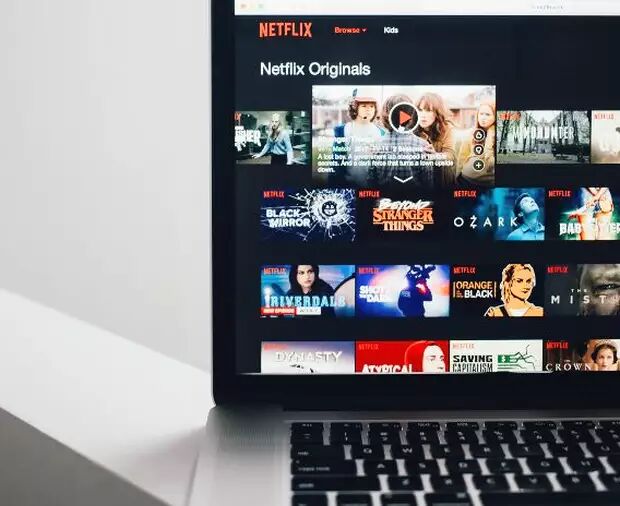A laptop displays the Netflix homepage, showcasing a variety of original content like "Black Mirror," "Stranger Things," and "Ozark." The interface is clean and inviting.
