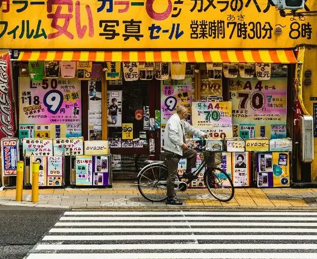 A man with a bicycle stands in front of a Japanese store selling film, photo prints, and DVD copies. The windows are covered in special offers and prices.
