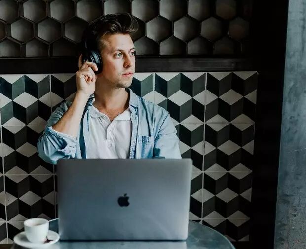 A young man wearing headphones sits in front of a laptop, looking thoughtfully out the window while listening to music.