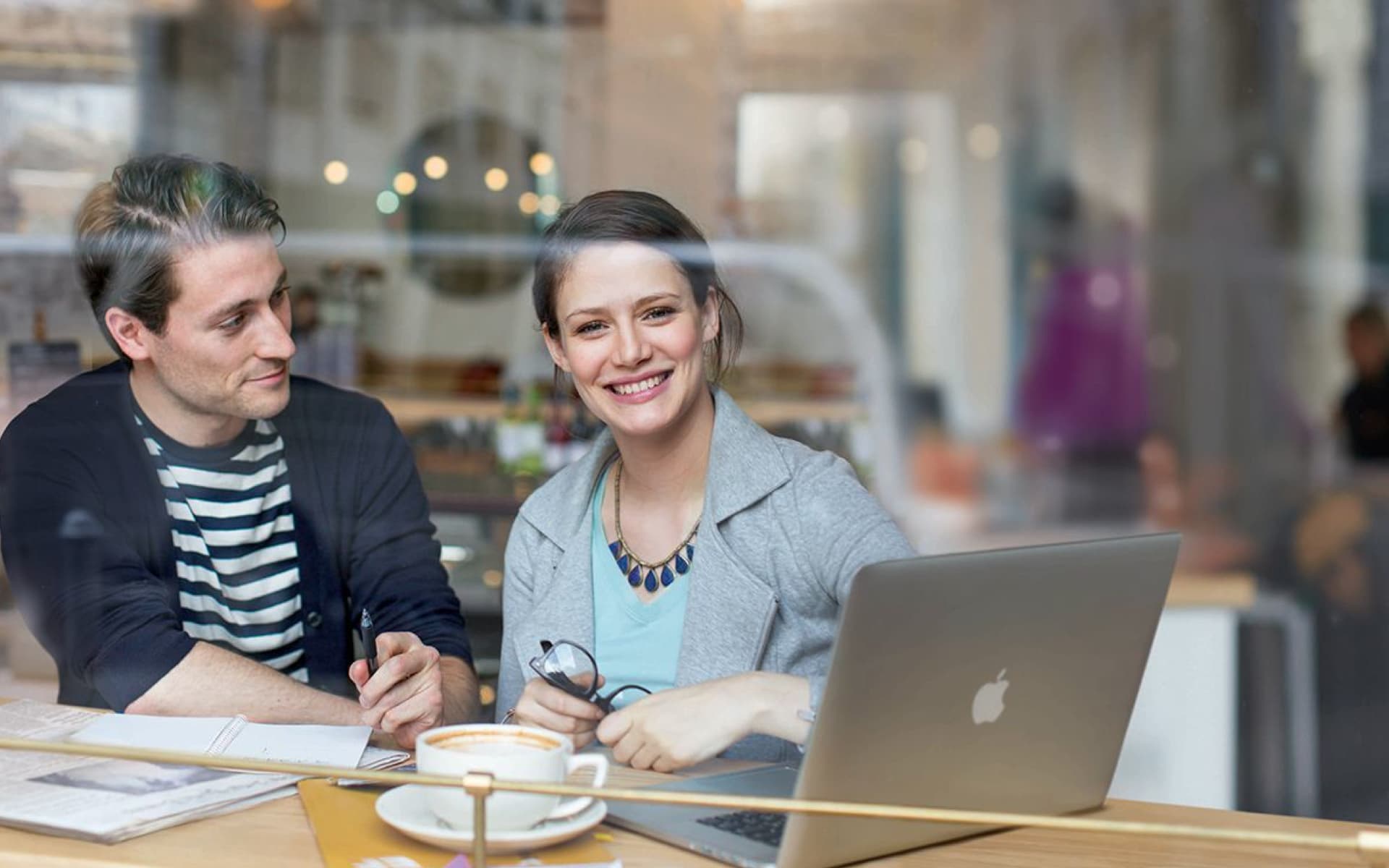 Two colleagues, a man and a woman, are sitting at a table in a cafe, looking at a laptop and smiling. They appear to be engaged in a work-related discussion.
