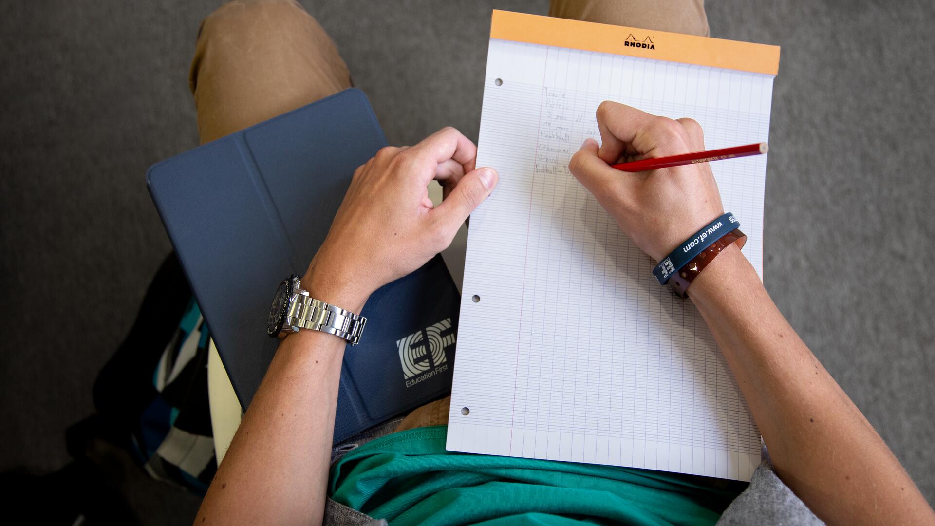 The image shows a person sitting down, holding a tablet on their lap with their left arm, and writing on a notepad with a red pencil in their right hand. The tablet has "EF" visible on its cover.