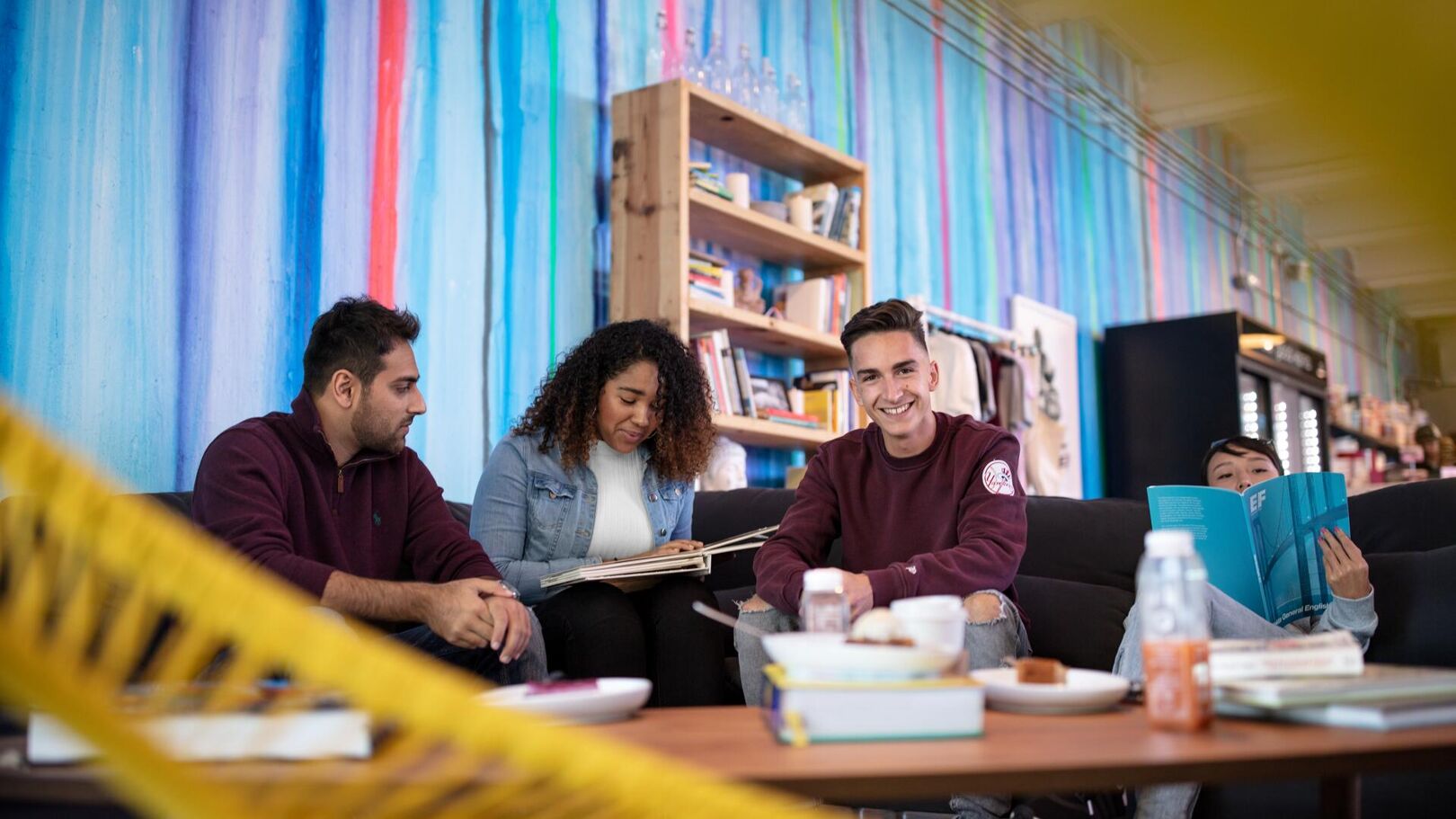 Four young adults are sitting on a couch in a bright room featuring a colorful mural and bookshelves. They appear to be socializing and studying together.