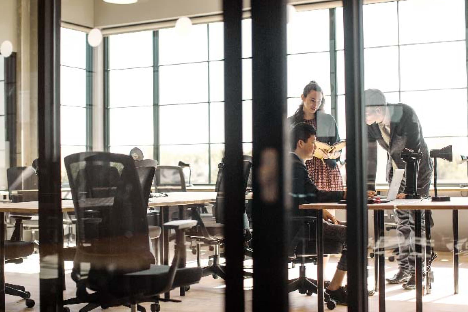 Three colleagues are collaborating around a desk in a modern office space with large windows, discussing a project on a laptop. The atmosphere appears professional and focused.