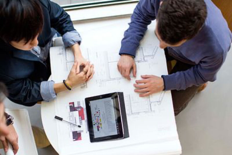 Two individuals are discussing blueprints and a tablet laid out on a table. They appear to be in an office or meeting setting, collaborating on a project.
