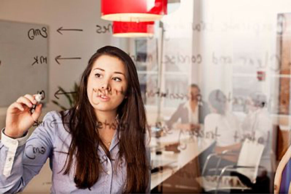 A woman is writing on a glass board in an office setting, with colleagues blurred in the background. The writing appears to be notes or a plan.