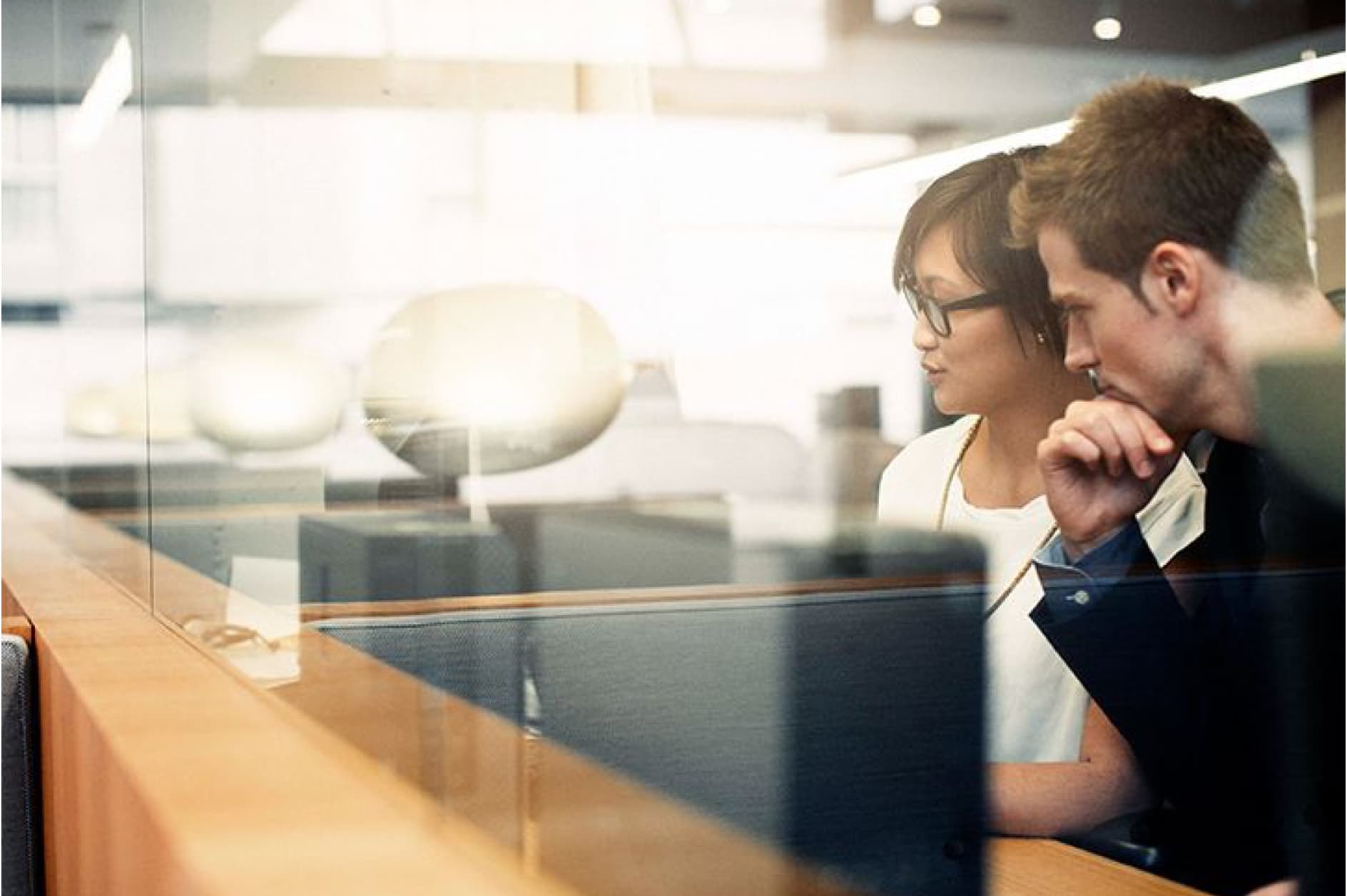 Two colleagues, one of East Asian descent and the other Caucasian, are intently focused on a computer screen in a modern office setting, possibly collaborating on a project.