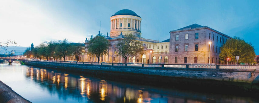 The image shows the Four Courts in Dublin at dusk, with the River Liffey flowing in front, reflecting the city lights.