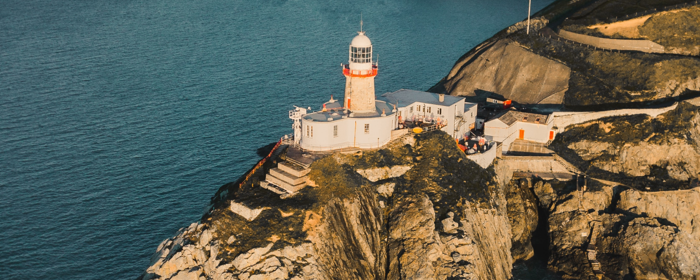 A lighthouse stands on a rocky cliff by the sea, with buildings and stairs leading to its entrance.