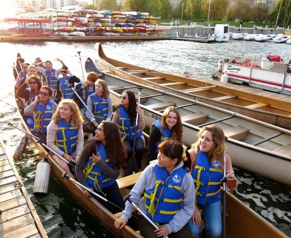 A group of women in a dragon boat holding paddles and traveling on the water, with kayaks visible in the background.