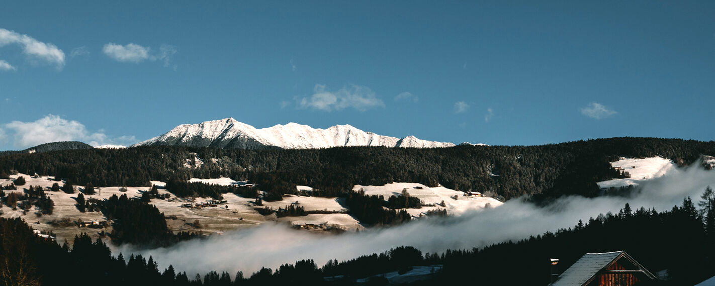 A serene winter landscape unfolds with snow-capped mountains in the distance, dark evergreen forests, and a small village nestled in the valley, partially obscured by mist.