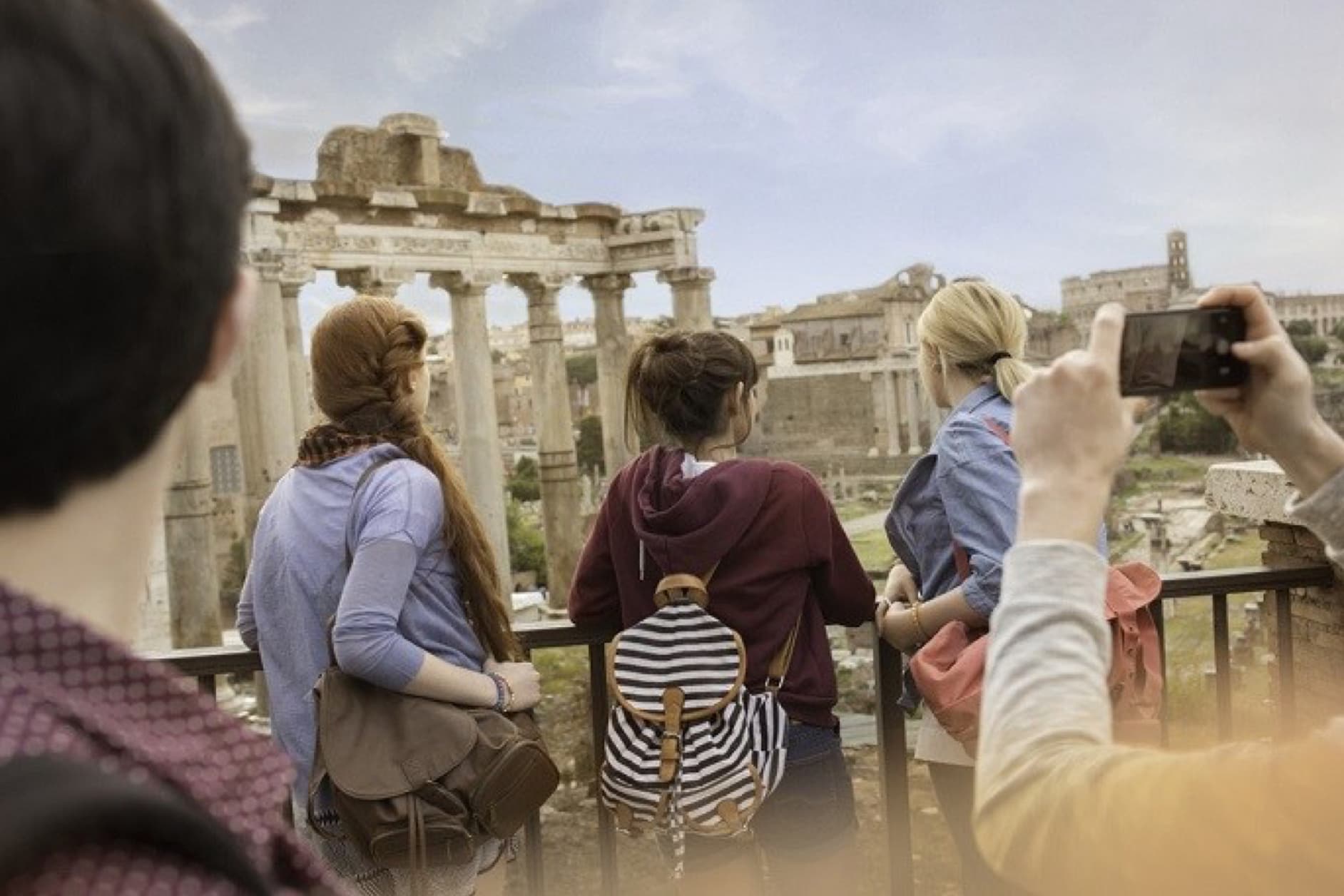 A group of tourists are admiring the ancient Roman Forum, with one person capturing the moment with their smartphone, showcasing a blend of historical exploration and modern technology.
