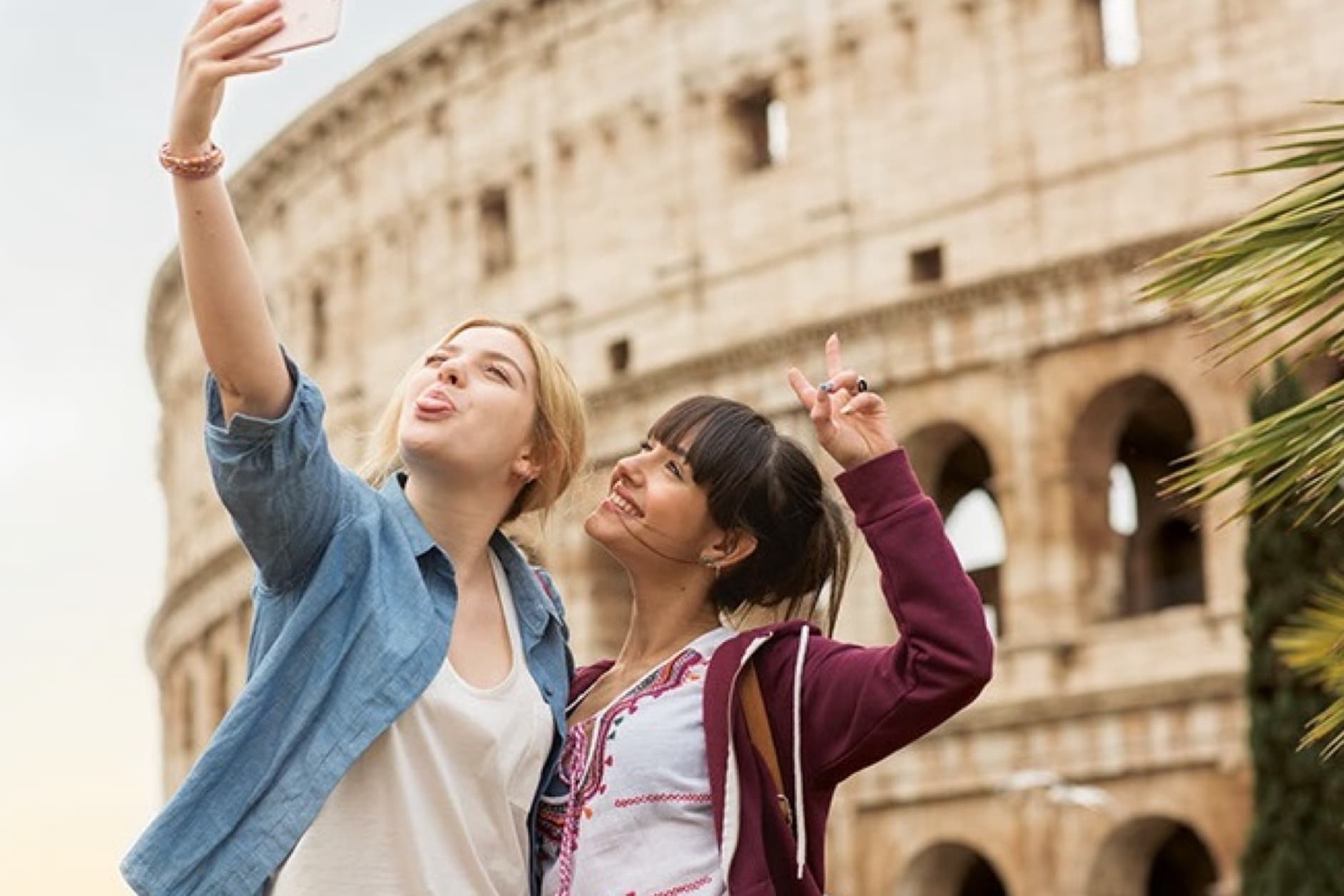 Two young women are taking a selfie with the Colosseum in Rome, Italy, in the background. They are smiling and making peace signs.