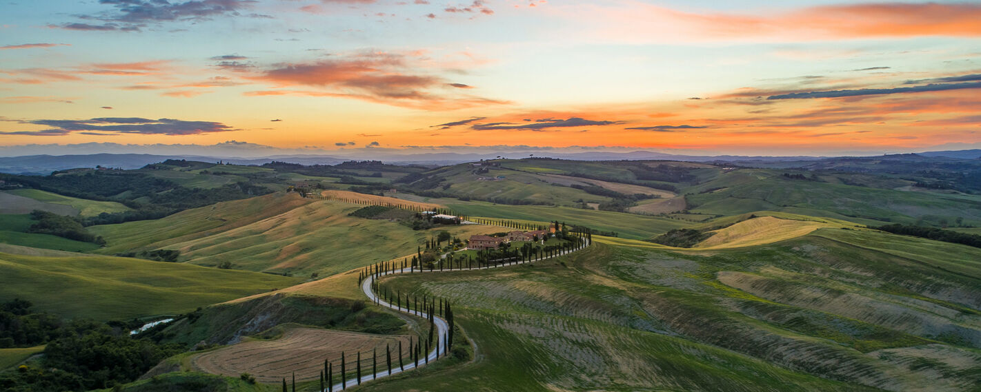 A winding road lined with cypress trees leads to a farmhouse nestled in the rolling hills of Tuscany during a vibrant sunset.