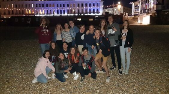 A group of about 15 young people are standing and sitting on a pebble beach in front of a lit-up building at night.