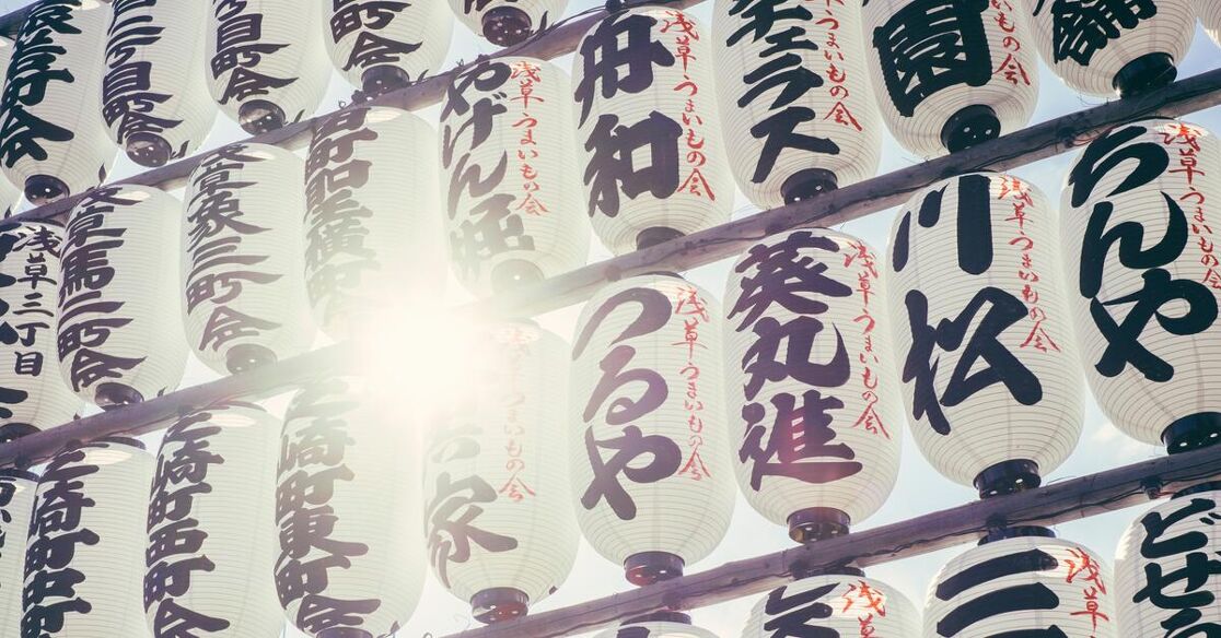 A row of white Japanese lanterns with black Kanji characters, held up by wooden beams, with sunlight shining through them.