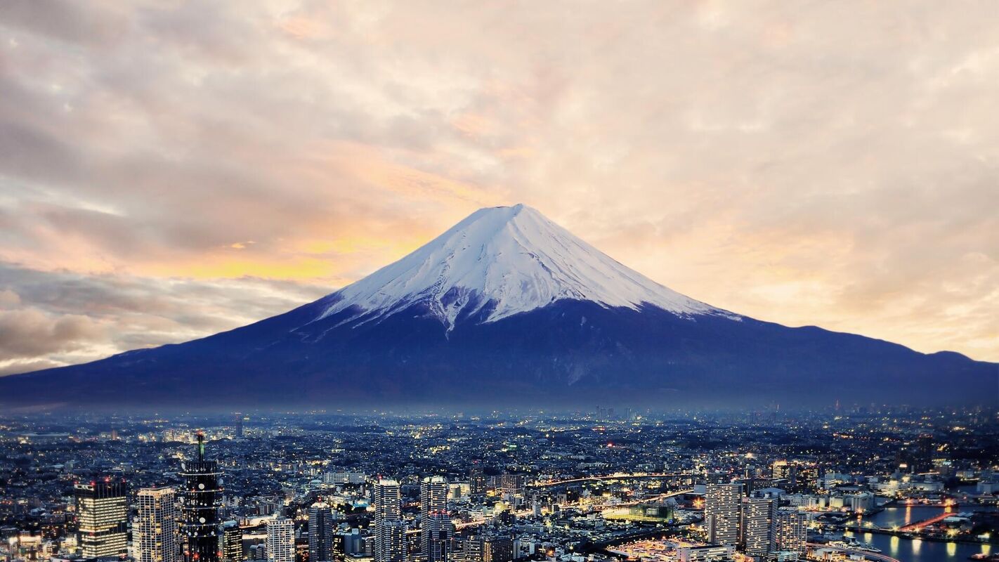 This image shows Mount Fuji, a majestic snow-capped volcano, towering over a sprawling cityscape at dusk. The sky is filled with warm, cloudy hues, creating a dramatic backdrop for the iconic natural landmark.