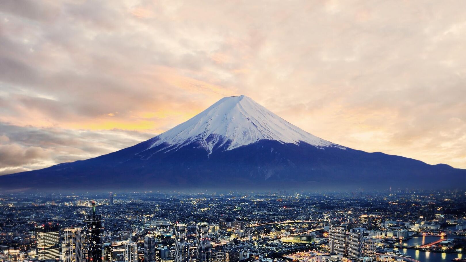 This image shows Mount Fuji, a majestic snow-capped volcano, towering over a sprawling cityscape at dusk. The sky is filled with warm, cloudy hues, creating a dramatic backdrop for the iconic natural landmark.