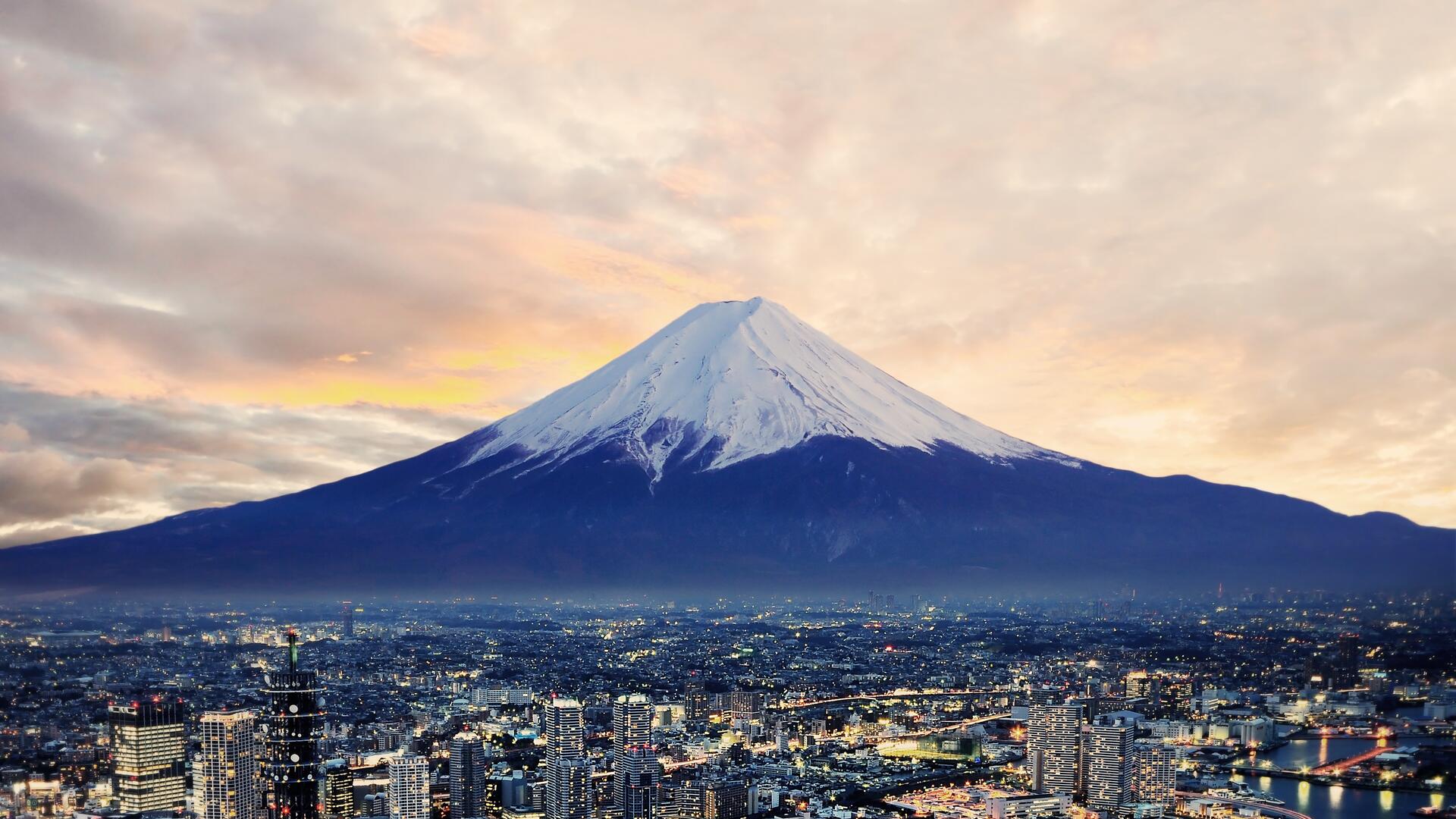 This image shows Mount Fuji, a majestic snow-capped volcano, towering over a sprawling cityscape at dusk. The sky is filled with warm, cloudy hues, creating a dramatic backdrop for the iconic natural landmark.