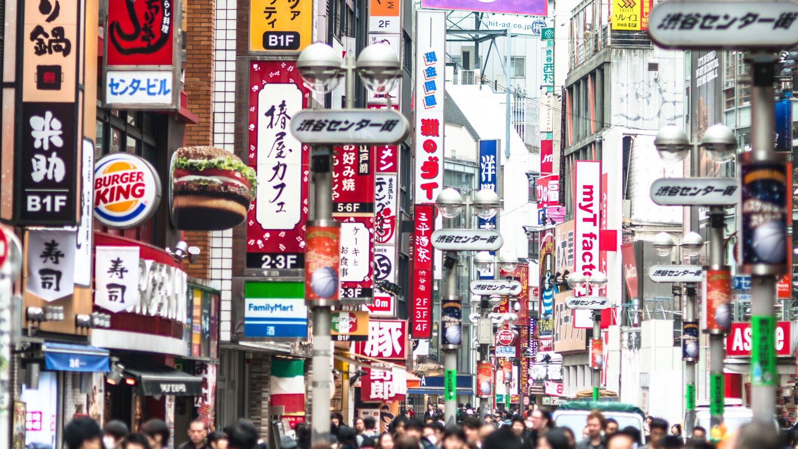 This image depicts a bustling street scene in Shibuya, Tokyo, Japan, characterized by numerous colorful signs and advertisements, including a Burger King, for various businesses and shops, creating a vibrant urban atmosphere.