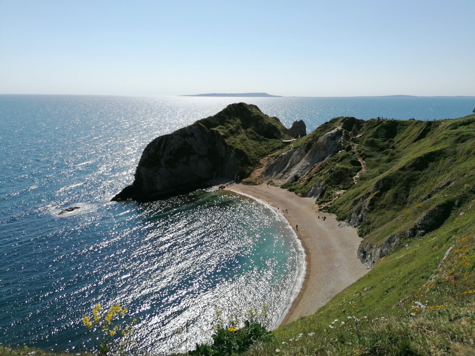 A scenic view of a crescent-shaped beach nestled between green, rolling hills and the sparkling blue ocean. The sun shines brightly, illuminating the sandy shore and the clear turquoise water.