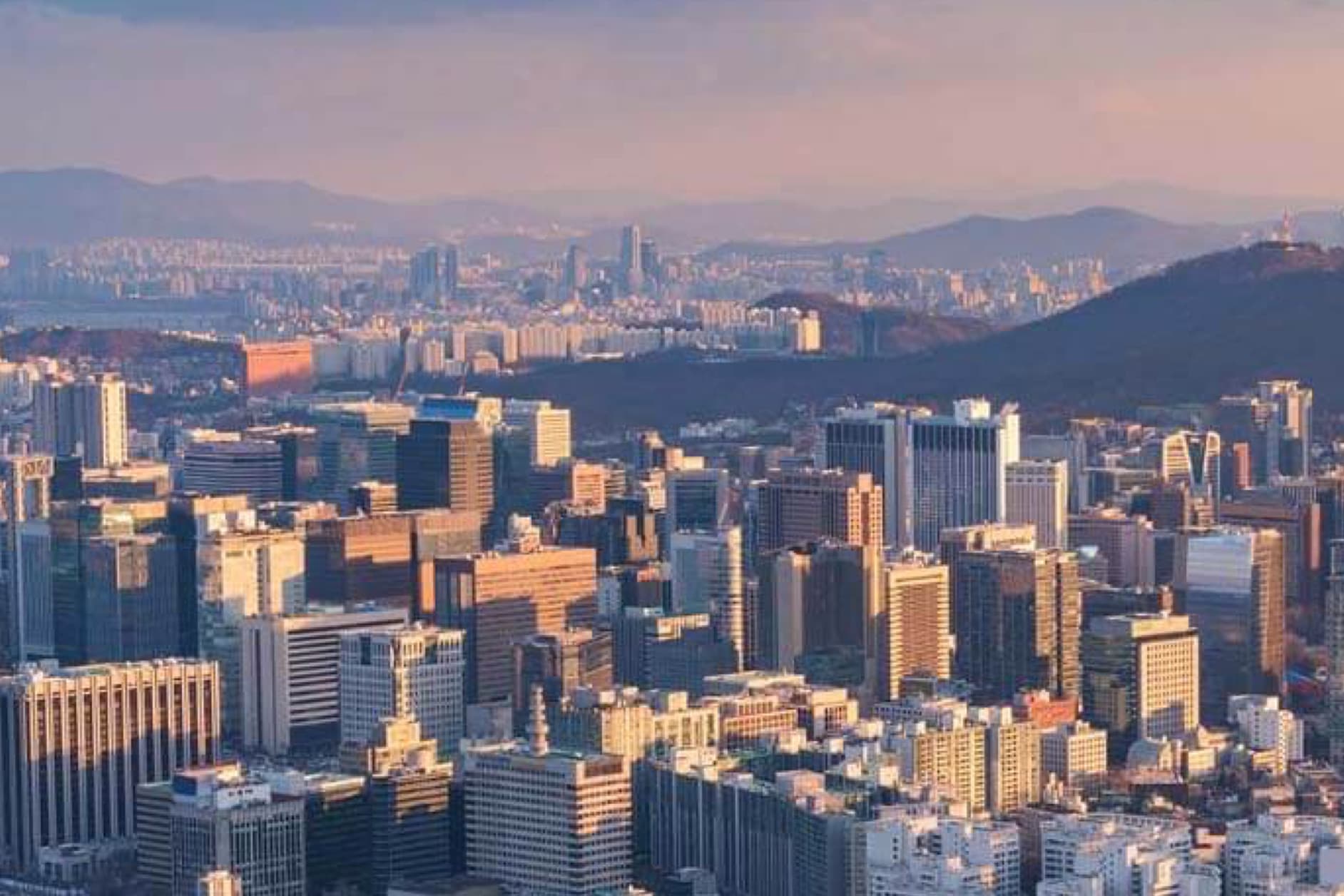The image displays a sprawling cityscape featuring numerous skyscrapers, with mountains visible in the background, all bathed in warm sunlight.