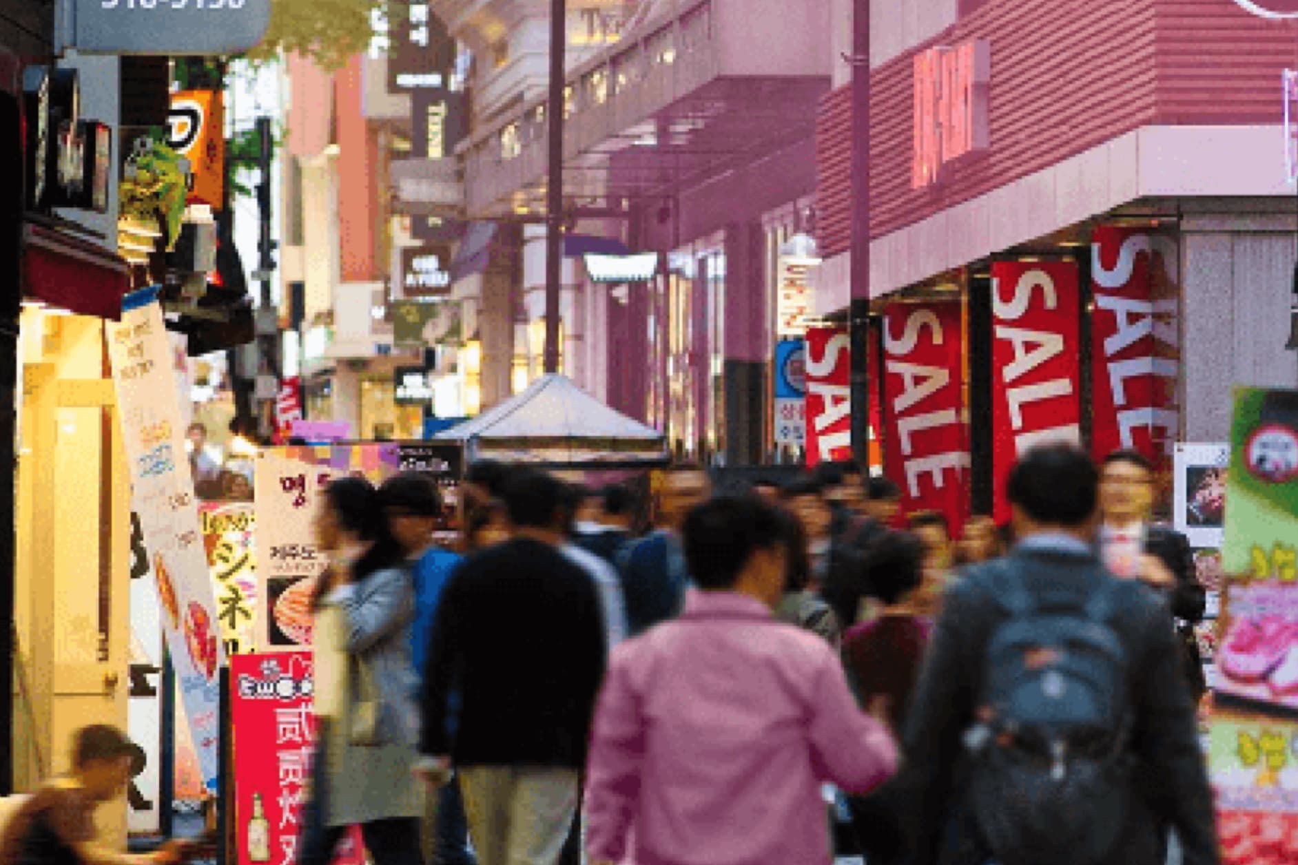 A bustling shopping street filled with people, shops, and sale banners. The architecture suggests a modern Asian city, vibrant with commercial activity.