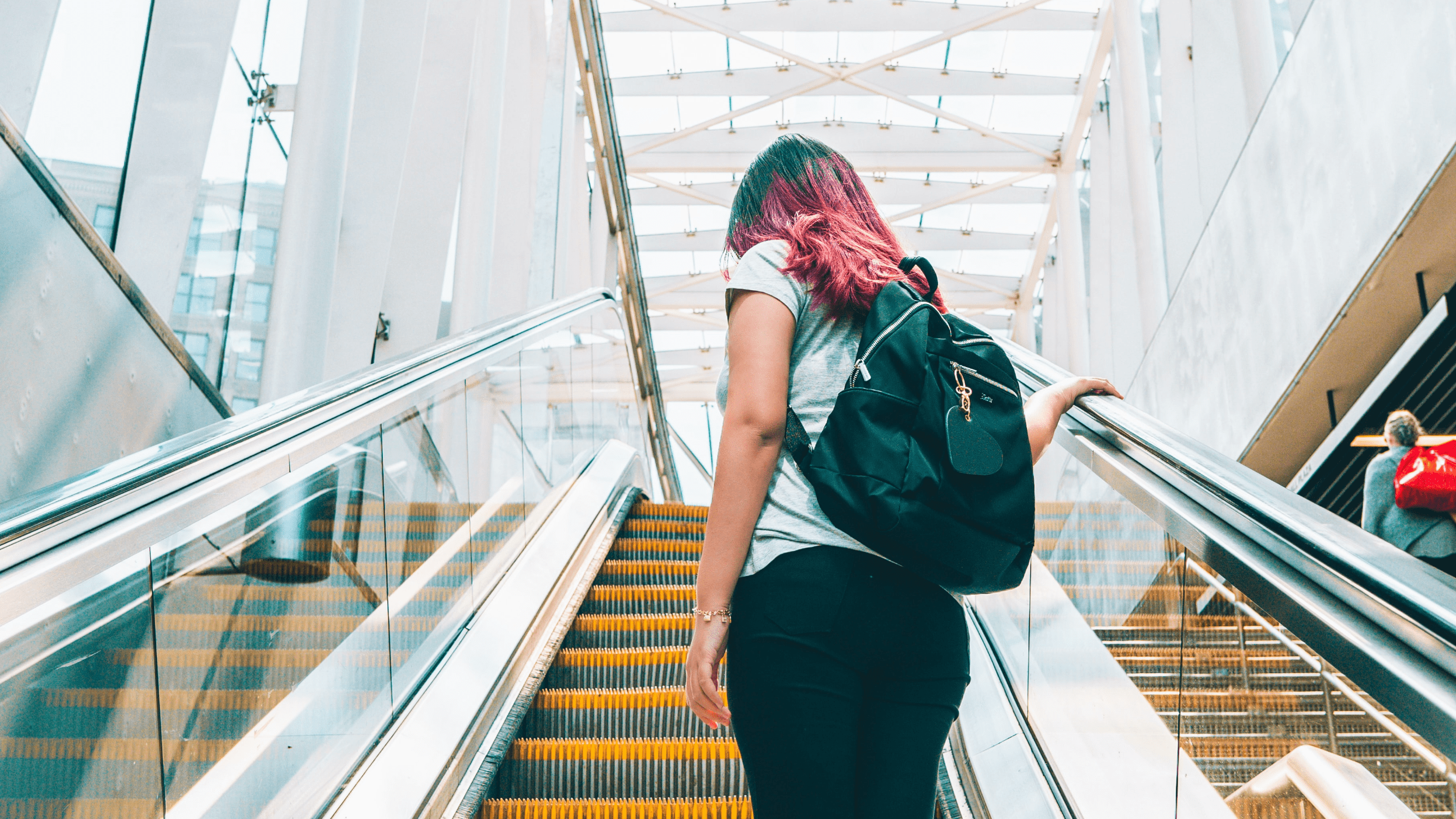 A person with pink and purple hair is riding an escalator upwards, carrying a black backpack. The escalator has yellow accents on the steps and is enclosed by modern architecture with glass and metal.