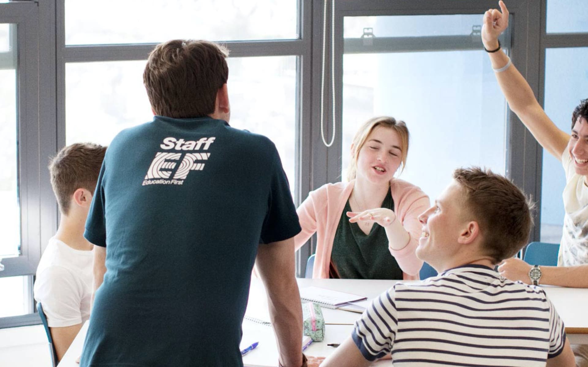 The image shows a group of young students and a staff member engaged in a discussion around a table, possibly in a classroom or workshop setting.