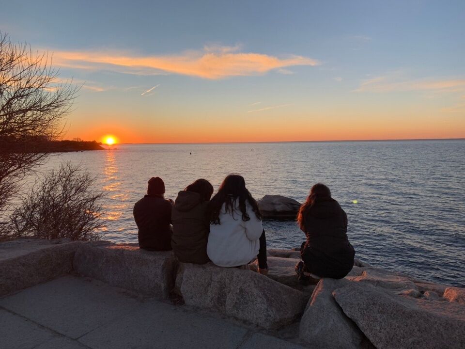 Four people sit on a rocky ledge, facing the ocean as the sun sets, casting a warm glow across the water and sky.