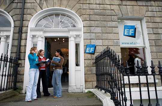Three young women stand outside an EF Education First language school building, engaged in conversation.