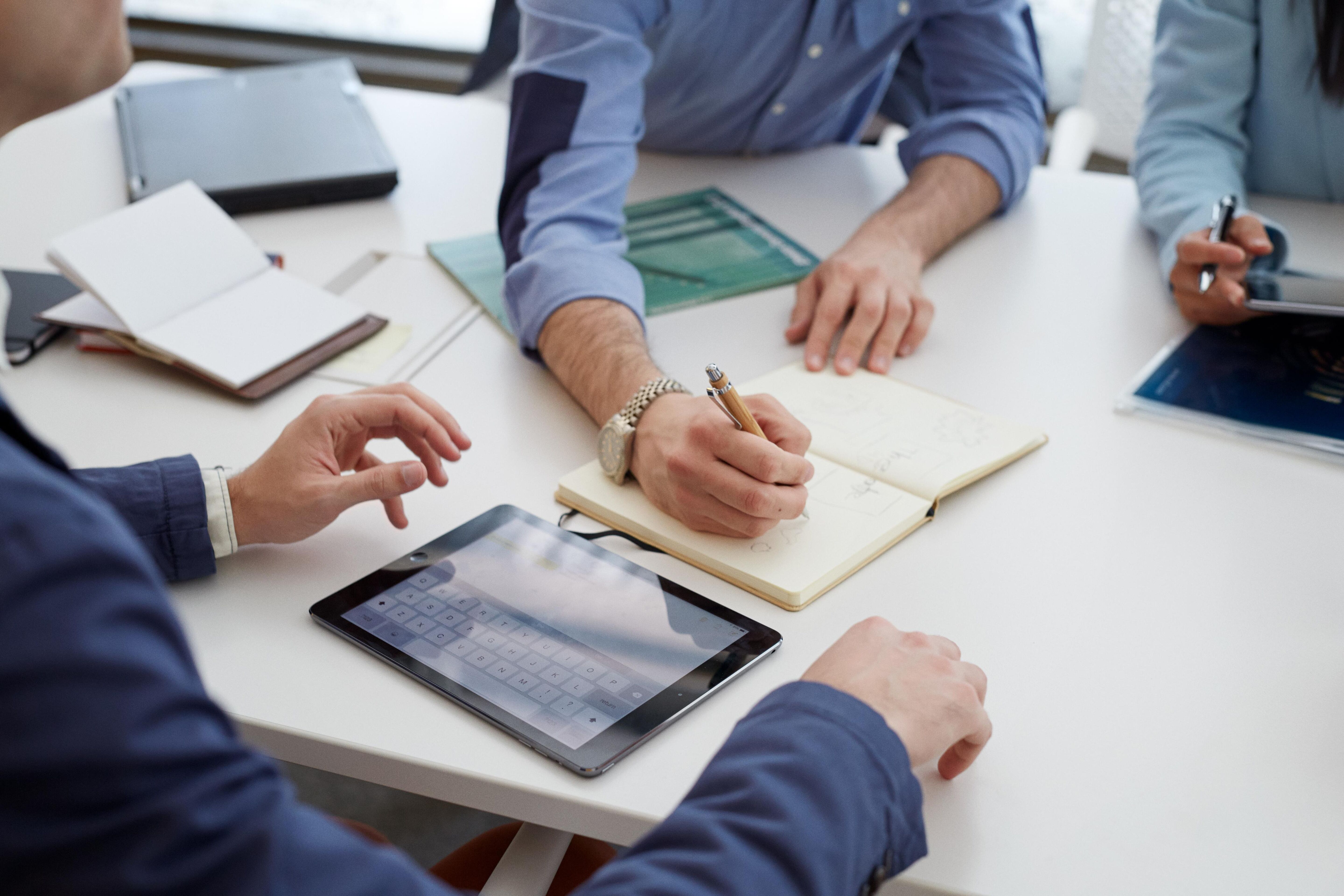 A group of business professionals gathered around a table for a meeting, with some taking notes and others looking at tablets.