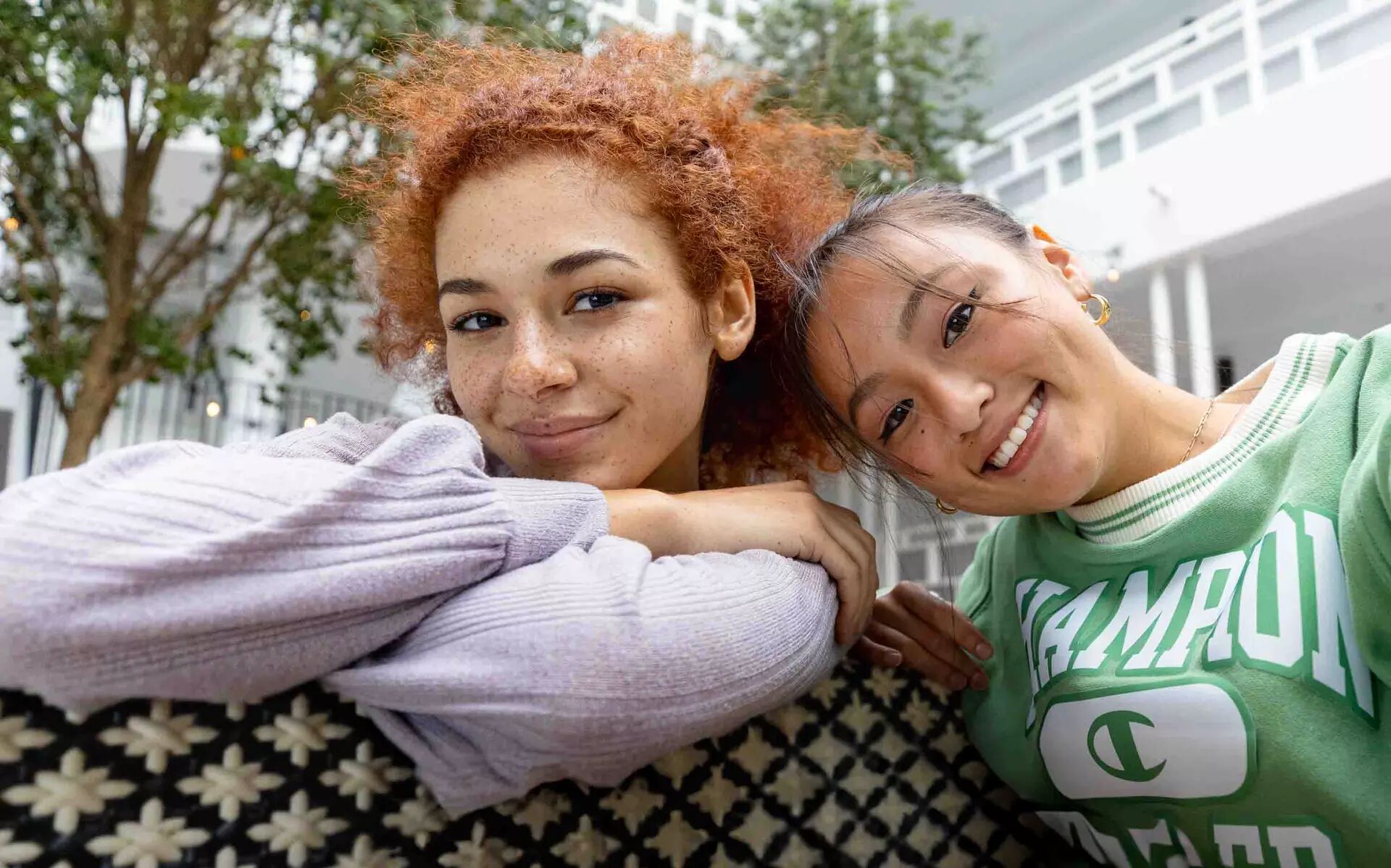 Two young women, one with red hair and the other with dark hair, smile at the camera while relaxing on a couch. They are dressed casually and appear to be enjoying each other's company.