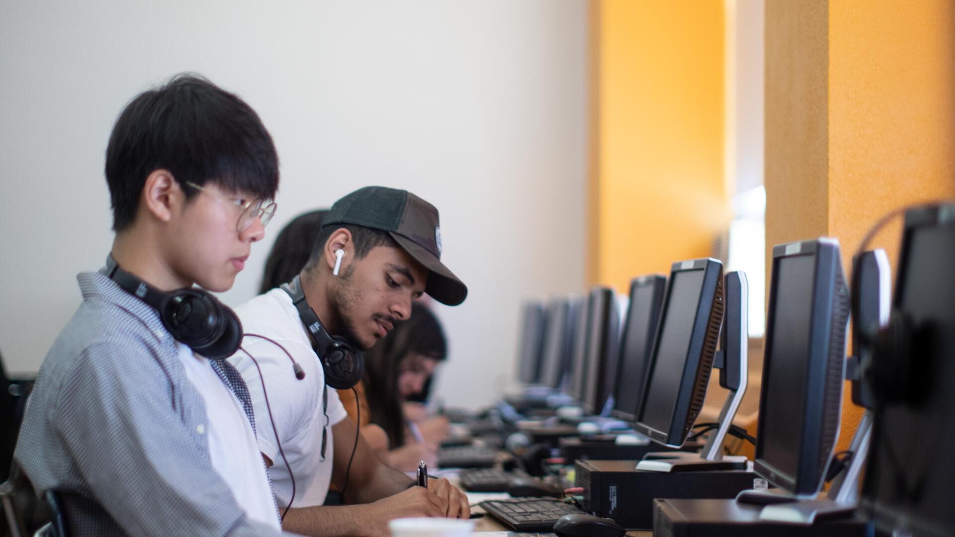 A group of young people are seated in a row, using computers. Some are wearing headphones, suggesting a focus on individual tasks or learning activities.