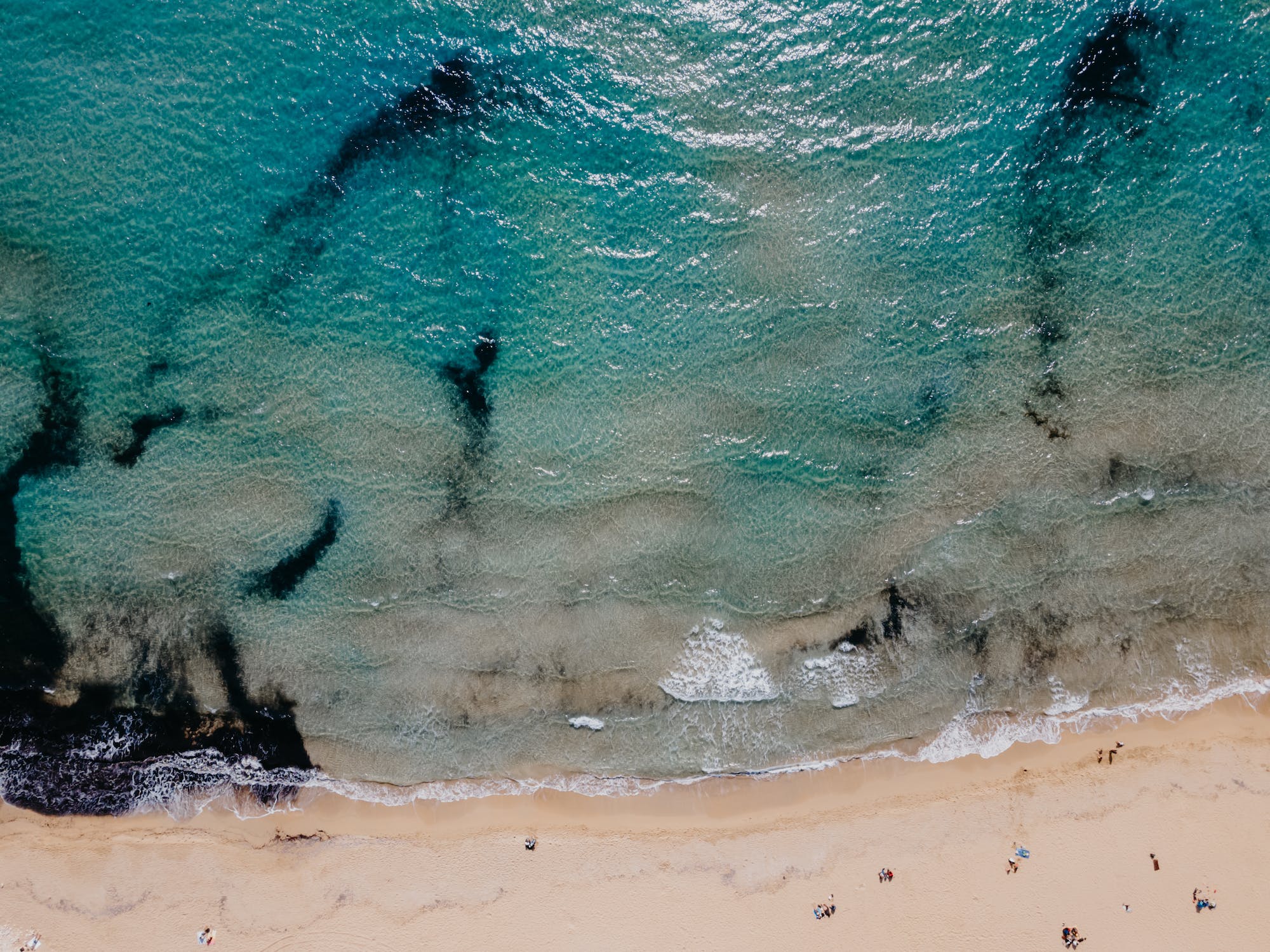 An aerial view shows a sandy beach meeting the clear turquoise water. Gentle waves lap the shore, and dark underwater rocks create abstract patterns beneath the surface.