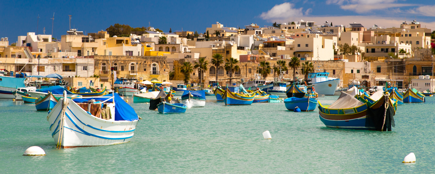 A picturesque harbor filled with traditional Maltese fishing boats, known as luzzus, painted in vibrant colors. The boats are moored in calm, turquoise waters with a backdrop of charming Mediterranean architecture.