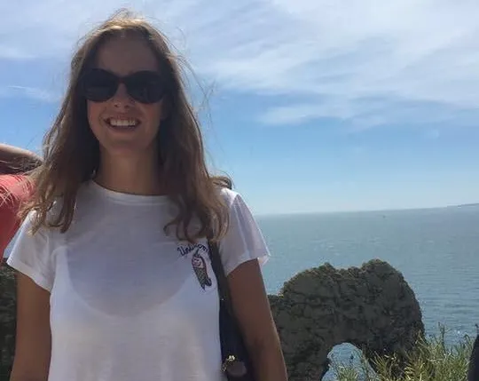 A young woman with long brown hair and sunglasses smiles at the camera, wearing a white t-shirt. She is outdoors with a scenic ocean view and a rock arch in the background.