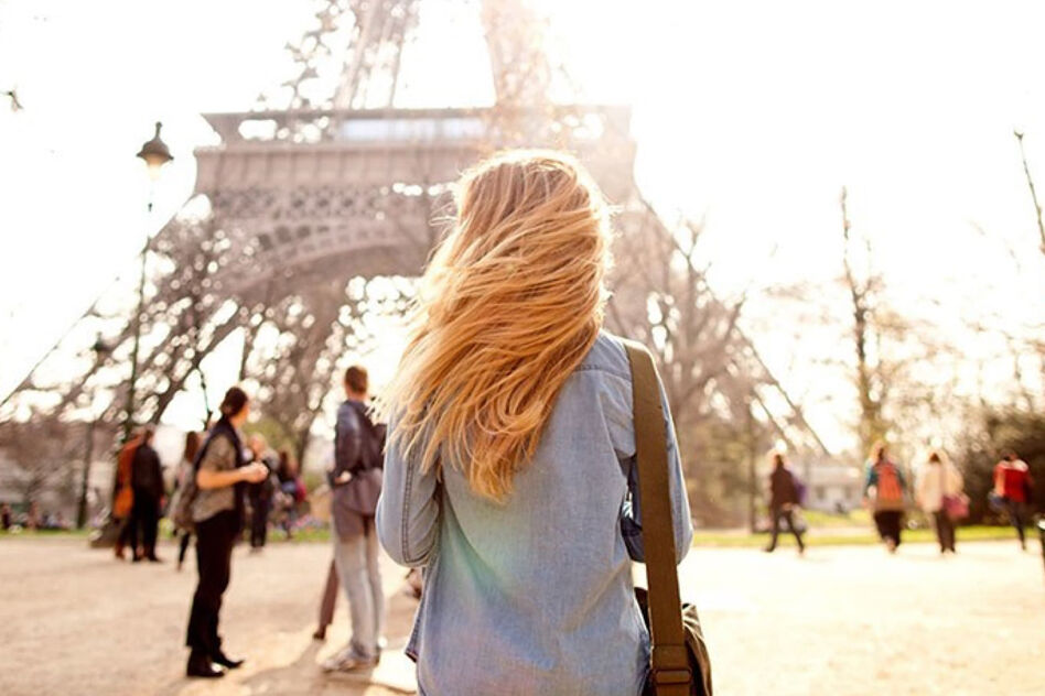 A young woman with long blonde hair stands with her back to the camera, looking towards the Eiffel Tower in Paris. The sun is shining brightly, casting a warm glow on the scene.