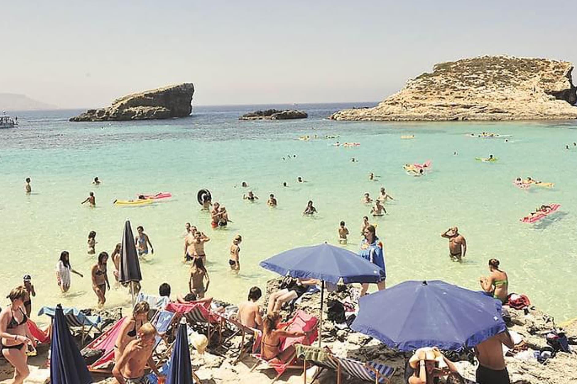 The image shows a crowded beach with many people swimming and sunbathing in the clear turquoise water. Rocky formations are visible in the sea under a bright, sunny sky.