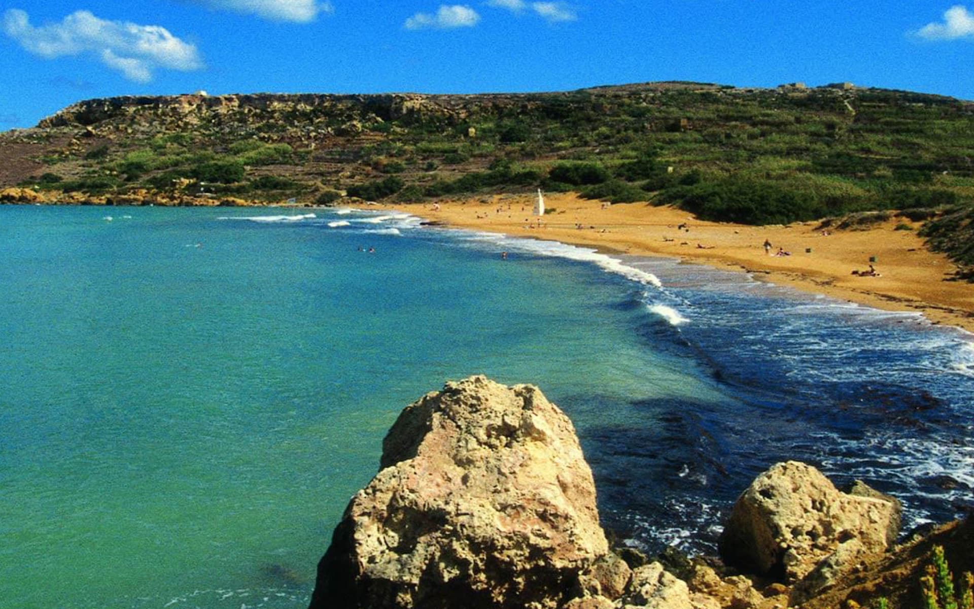 A picturesque beach with golden sand and turquoise waters, bordered by lush hills and rocky outcrops. People are enjoying the sun and sea at this idyllic vacation spot.